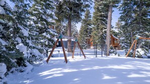 Un espacio exterior cubierto de nieve con columpios de madera y un área de juegos cercada, rodeado de altos árboles de hoja perenne.