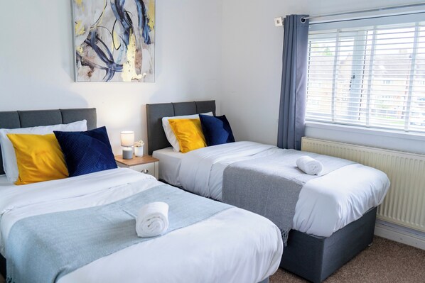 Ein Hotelzimmer mit zwei Betten, jeweils mit weißer und blauer Bettwäsche, einem Nachttisch mit Lampe und einem Fenster mit Jalousien.