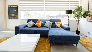A modern living room with a blue sectional sofa, a white coffee table, and a large window with blinds. - Apartment - Sleeps 6 - Parking - WiFi (Guildford)