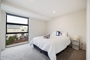 A modern bedroom with a large window, a bed with white bedding, a nightstand with a lamp, and a view of buildings outside. - Group and Family Friendly - 3 Bedroom Townhouse with Garage (Christchurch)