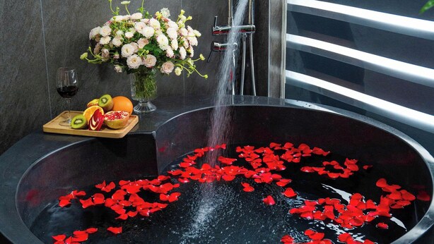 Une salle de bain moderne avec une grande baignoire remplie d’eau et de pétales de rose, un verre de vin et un plateau de fruits.