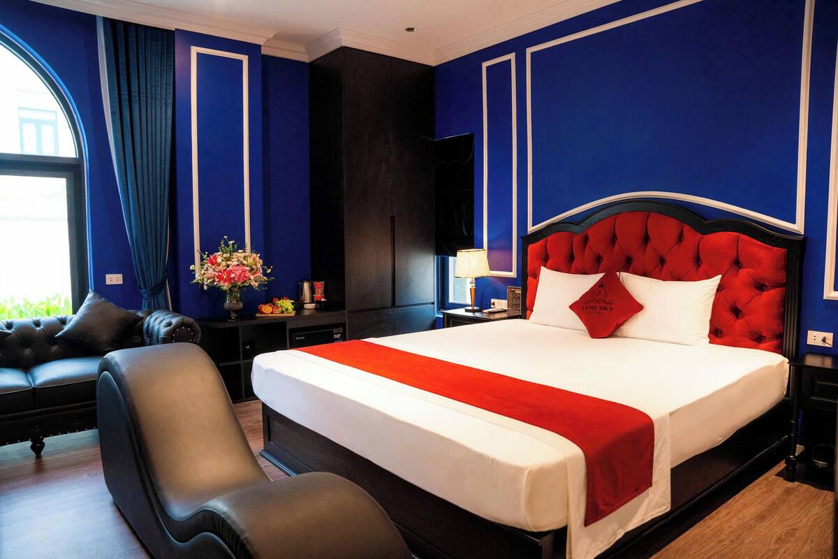 A hotel room with a large bed, a black leather chair, a desk with a lamp, and a large window with curtains.