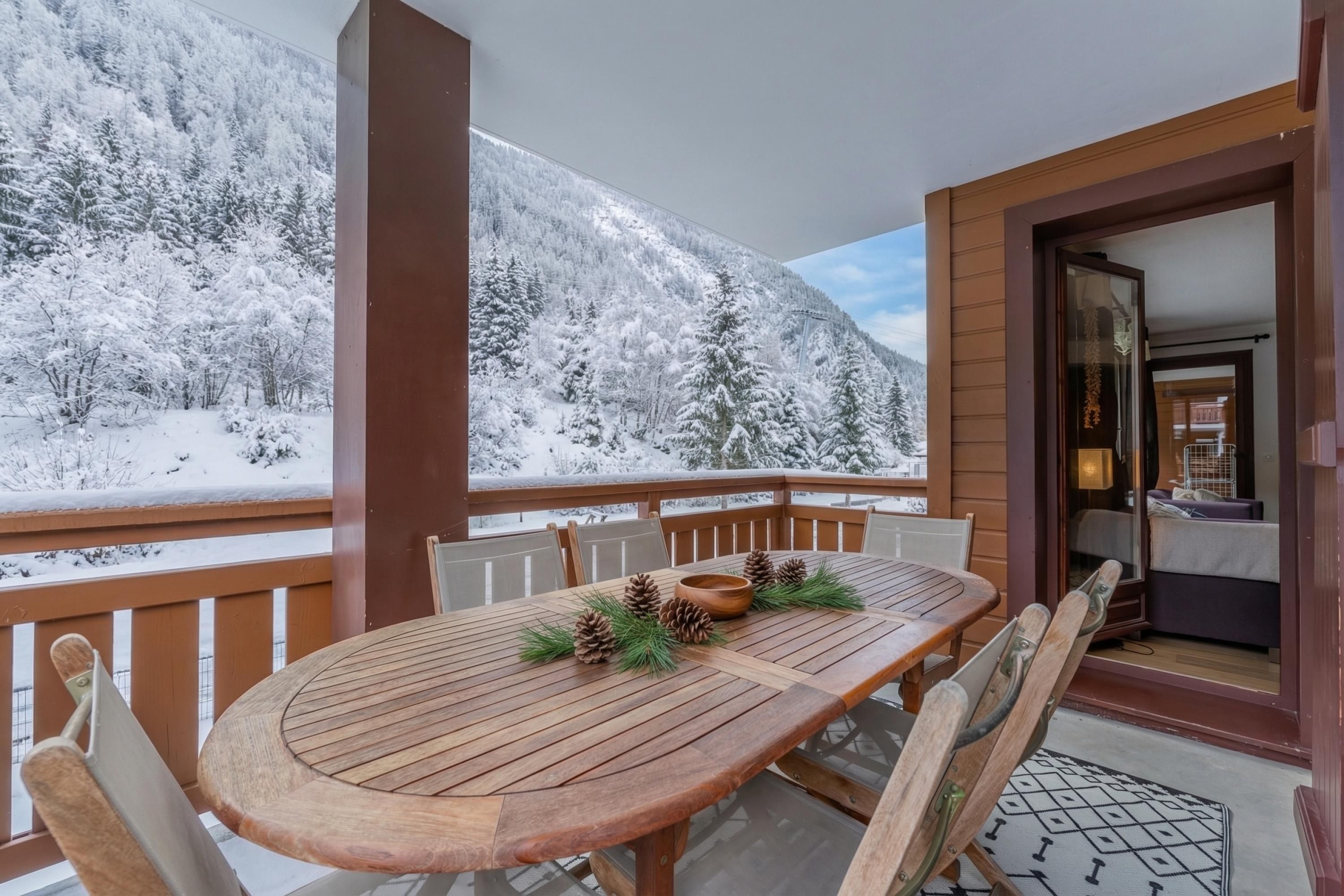 Une table en bois, entourée de chaises, sur un balcon, avec vue sur un paysage montagneux enneigé.
