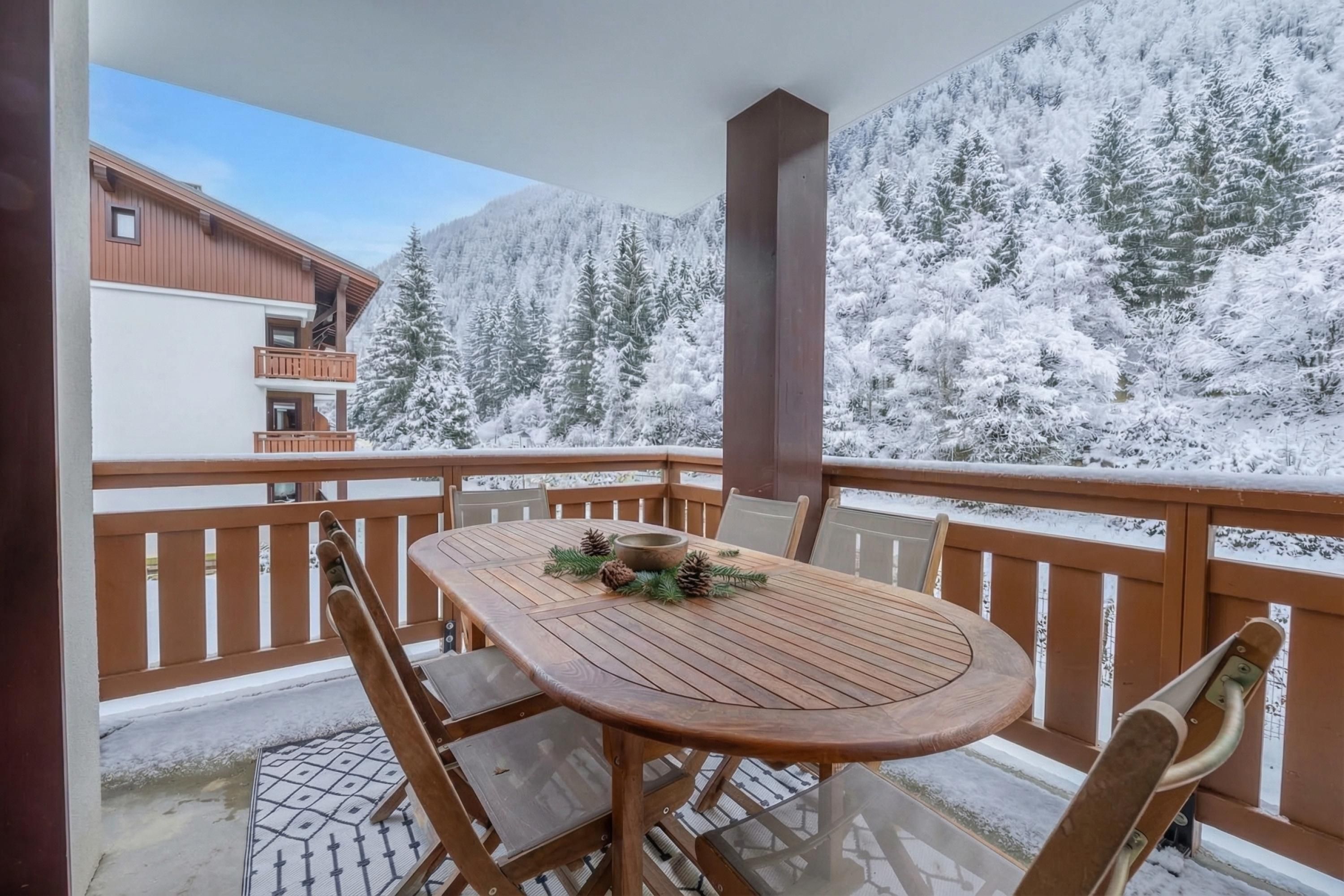 Una mesa de madera con sillas en un balcón con vistas a árboles nevados y montañas.