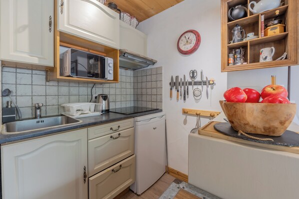 Une cuisine avec des meubles blancs, un four à micro-ondes, un grille-pain, un évier et un saladier en bois contenant des pommes, posé sur une planche à découper.