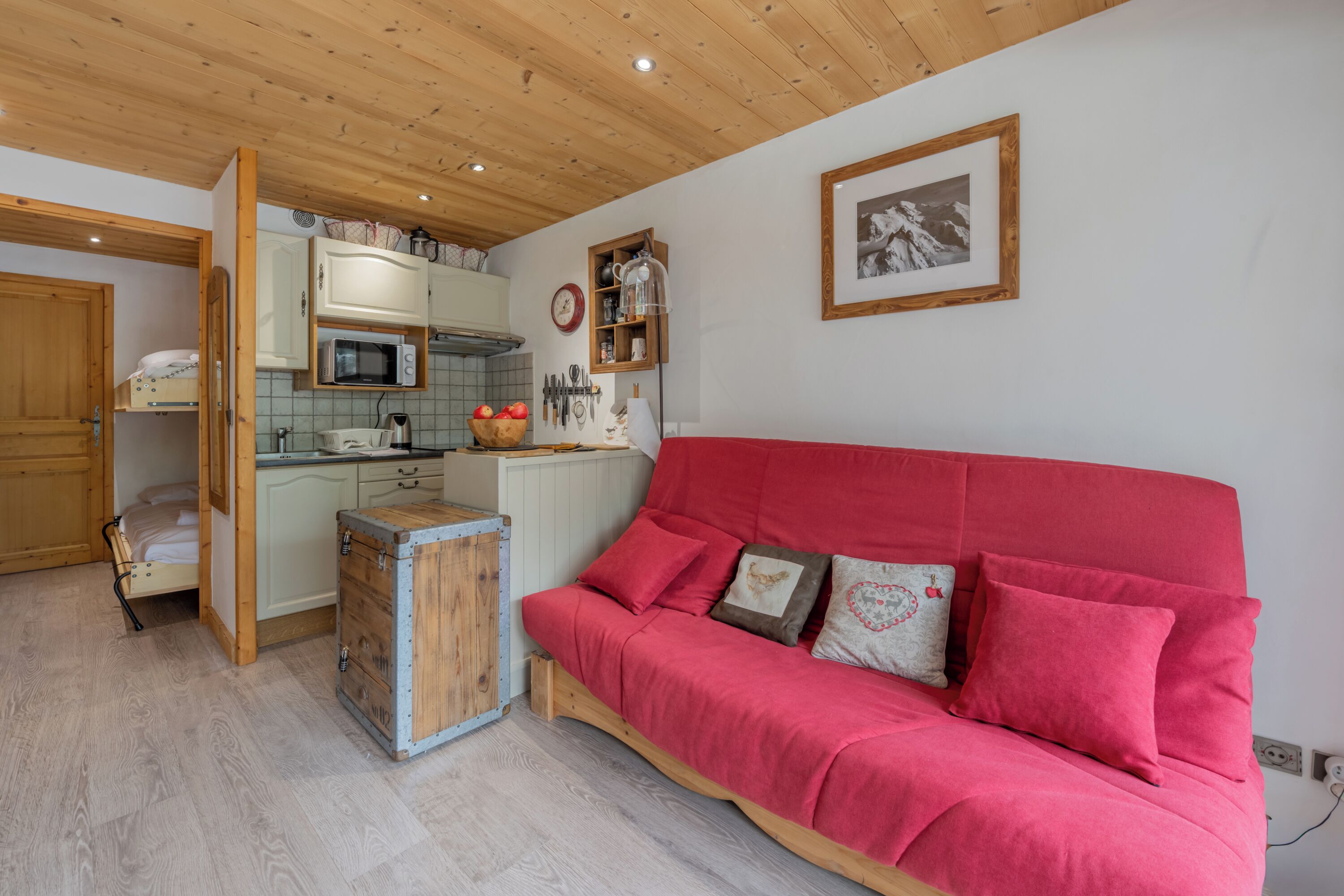 Una habitación acogedora con un sofá rojo, armarios de madera, una zona de cocina y un cuadro en la pared.