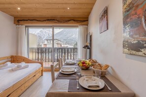 A room with a wooden bed, a dining table set with plates, glasses, and a bowl of fruit. - Clos Du Savoy - South Facing Balcony, Chamonix, France (Chamonix)