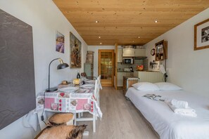 A cozy room with a wooden ceiling, a dining table with a checkered tablecloth, a bed with white linens, and a kitchen area with cabinets and a microwave.