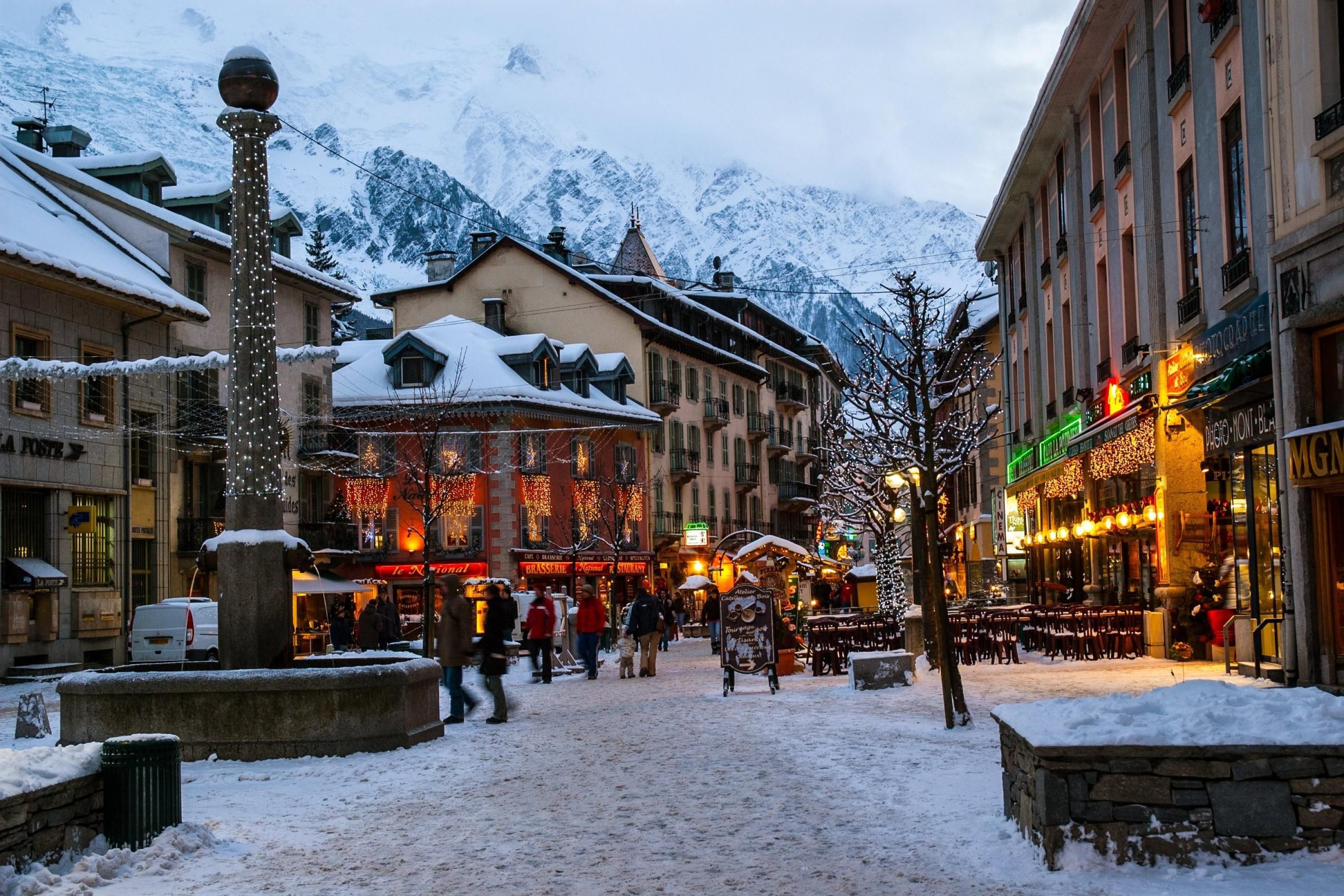 Une place enneigée, avec des bâtiments, une fontaine et une chaîne de montagnes en arrière-plan.