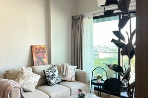 A living room with a sofa, a painting, a potted plant, and a view of a cityscape through a window.