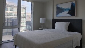 A hotel room with a large bed, a nightstand with a lamp, a painting of a forest, and a view of buildings through a large window.