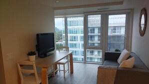 A modern living room with a sofa, a TV on a wooden table, and large windows offering a city view.