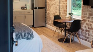 Ein gemütliches Schlafzimmer mit Bett, kleinem Tisch und Stuhl. Im Hintergrund ist eine Küchenzeile mit Kühlschrank und Mikrowelle zu sehen.