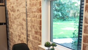 Ein Zimmer mit Backsteinwand, einem Fenster mit Jalousien, einem Holztisch, zwei schwarzen Stühlen und einem kleinen Blumentopf.