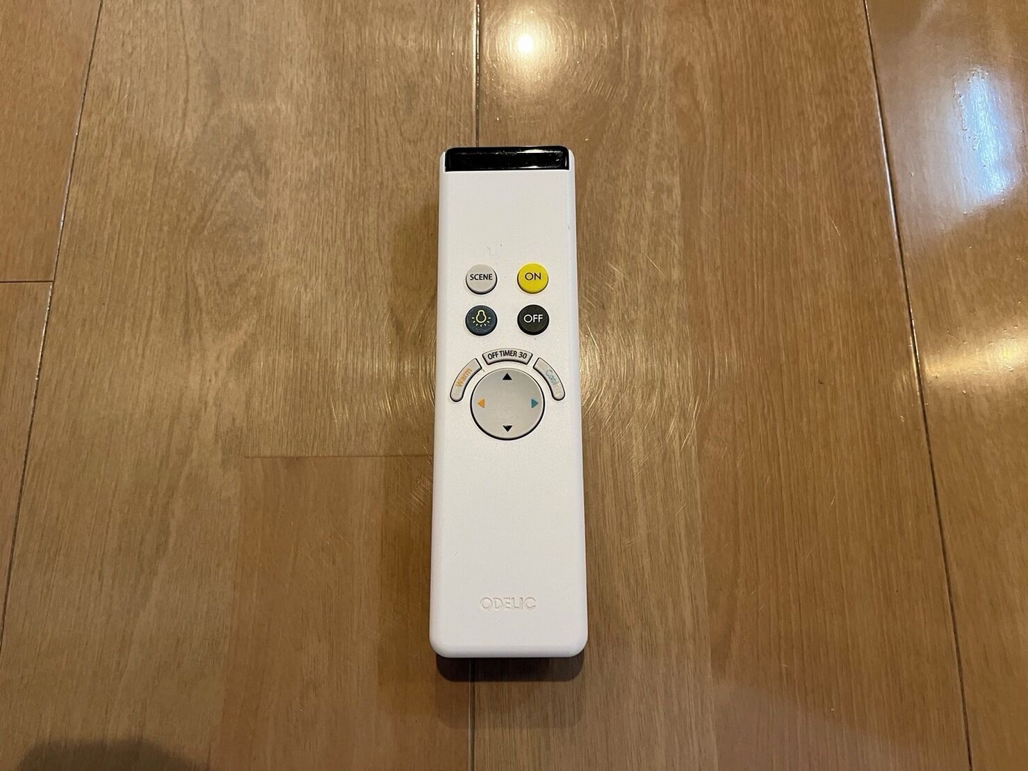 A white remote control with a circular navigation wheel, labeled 'ODERIC,' on a wooden floor.