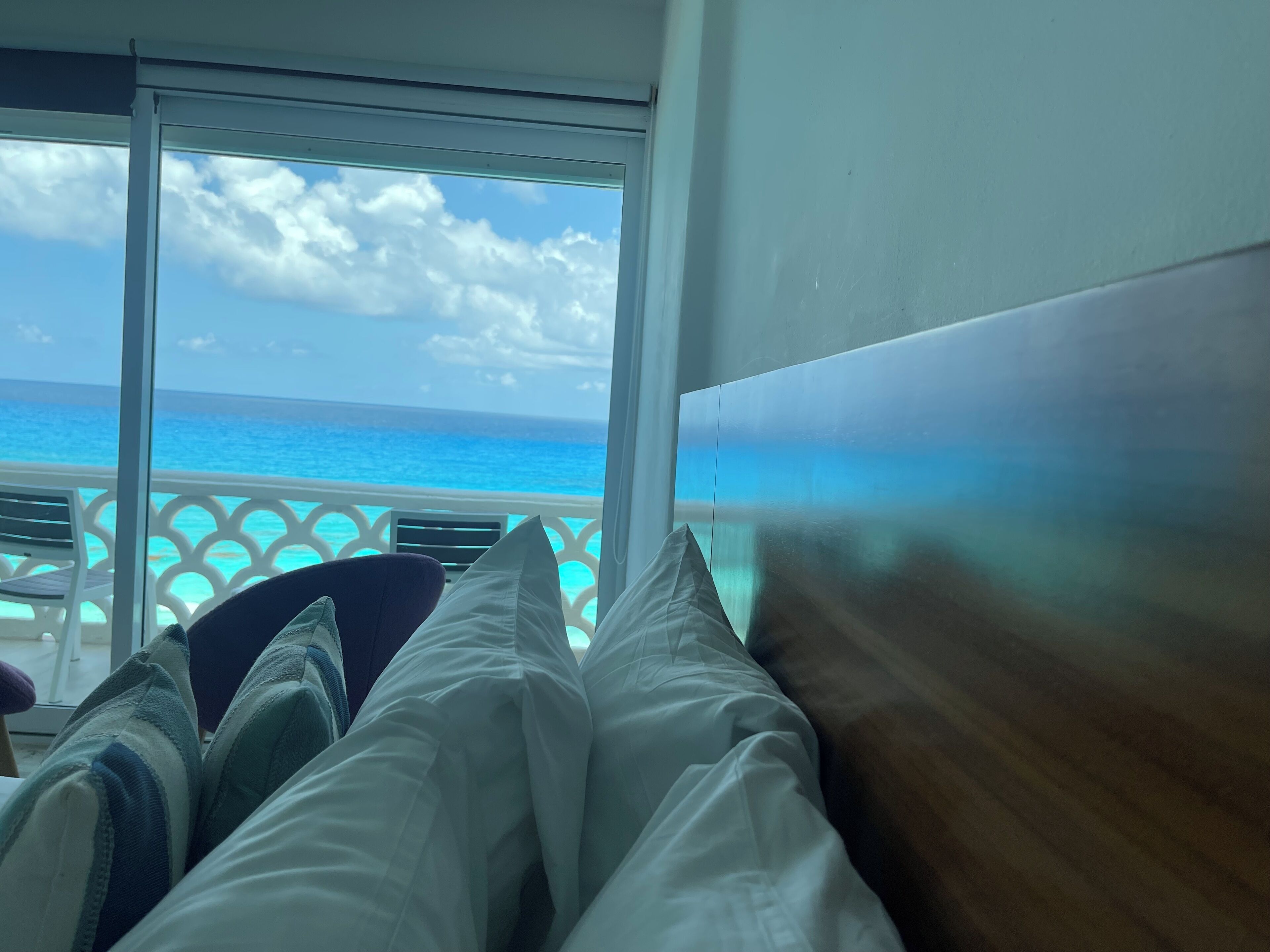 A room with a view of the ocean through large windows, a bed with white and striped bedding, and a wooden headboard.