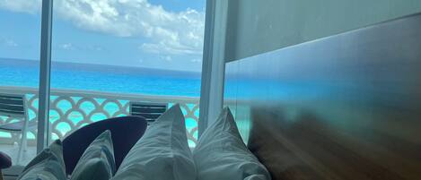 A room with a view of the ocean through large windows, a bed with white and striped bedding, and a wooden headboard.