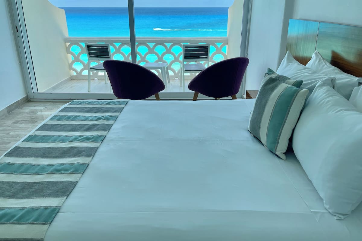 Ein Hotelzimmer mit einem großen Bett, zwei violetten Sesseln und Blick auf den Ozean vom Balkon.