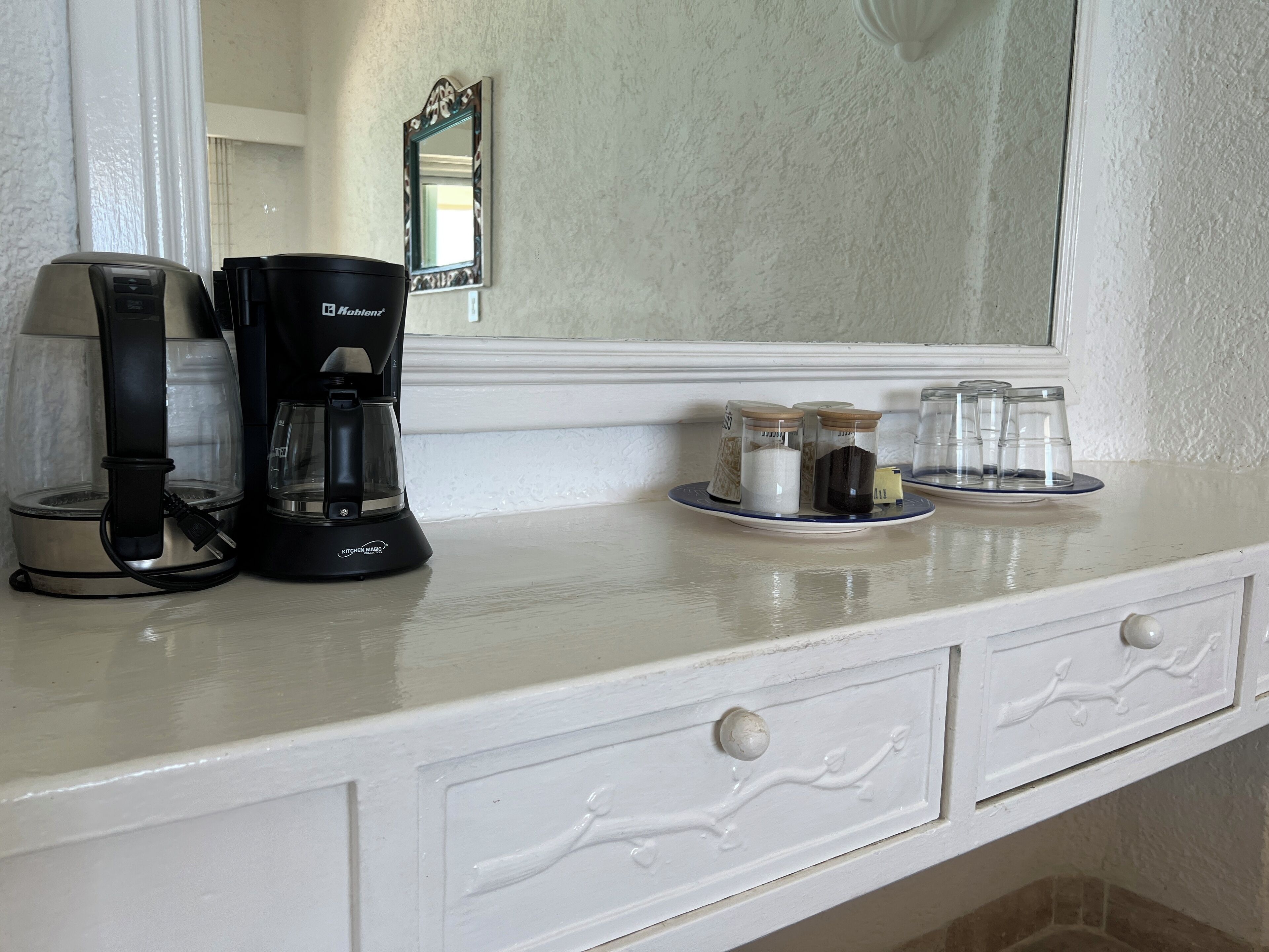 A kitchen counter with a coffee maker, a mirror, and glass jars on a plate.