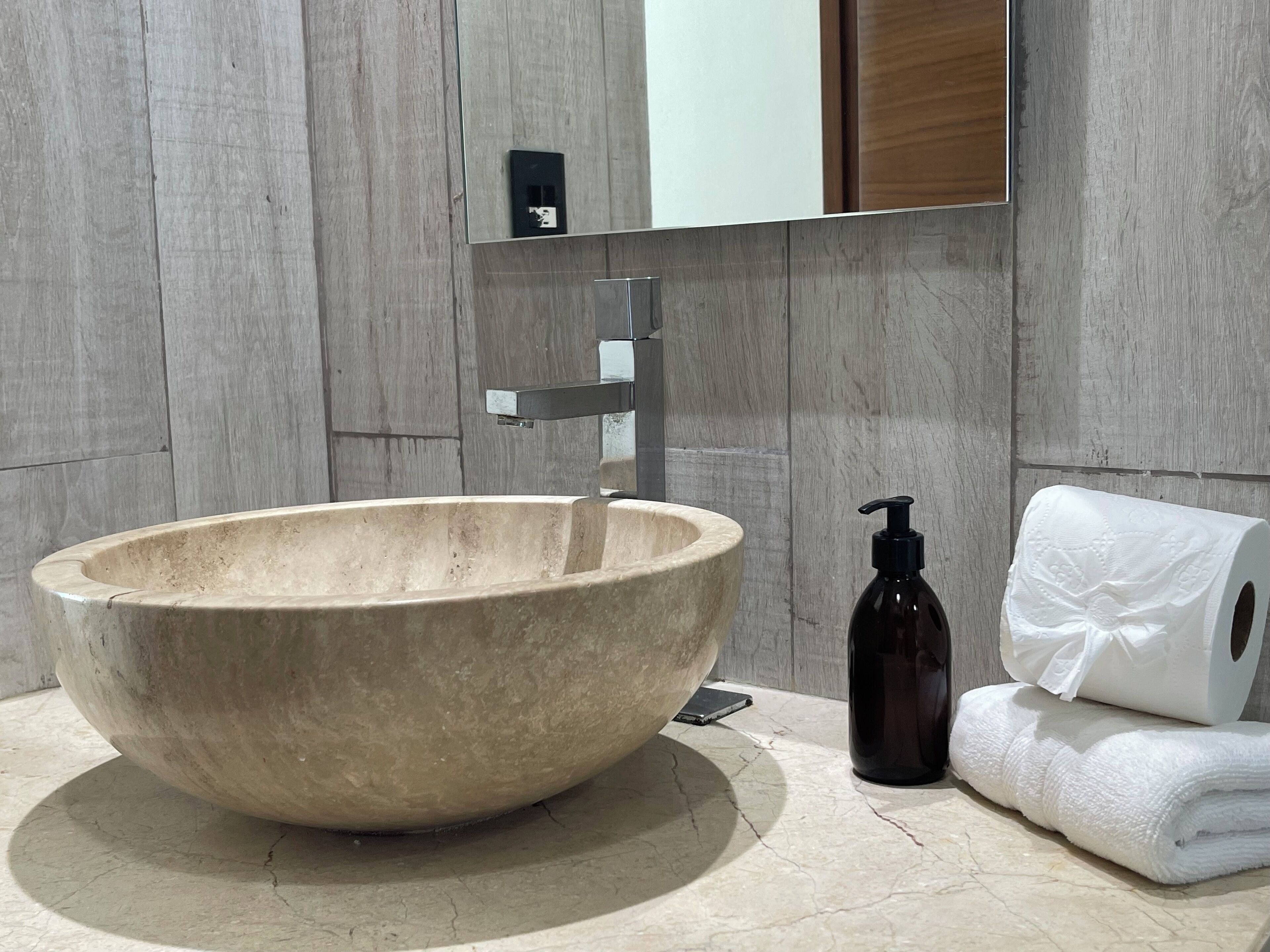 A large, round, beige ceramic sink with a modern faucet, a dark brown soap dispenser, and a roll of white toilet paper on a marble countertop.