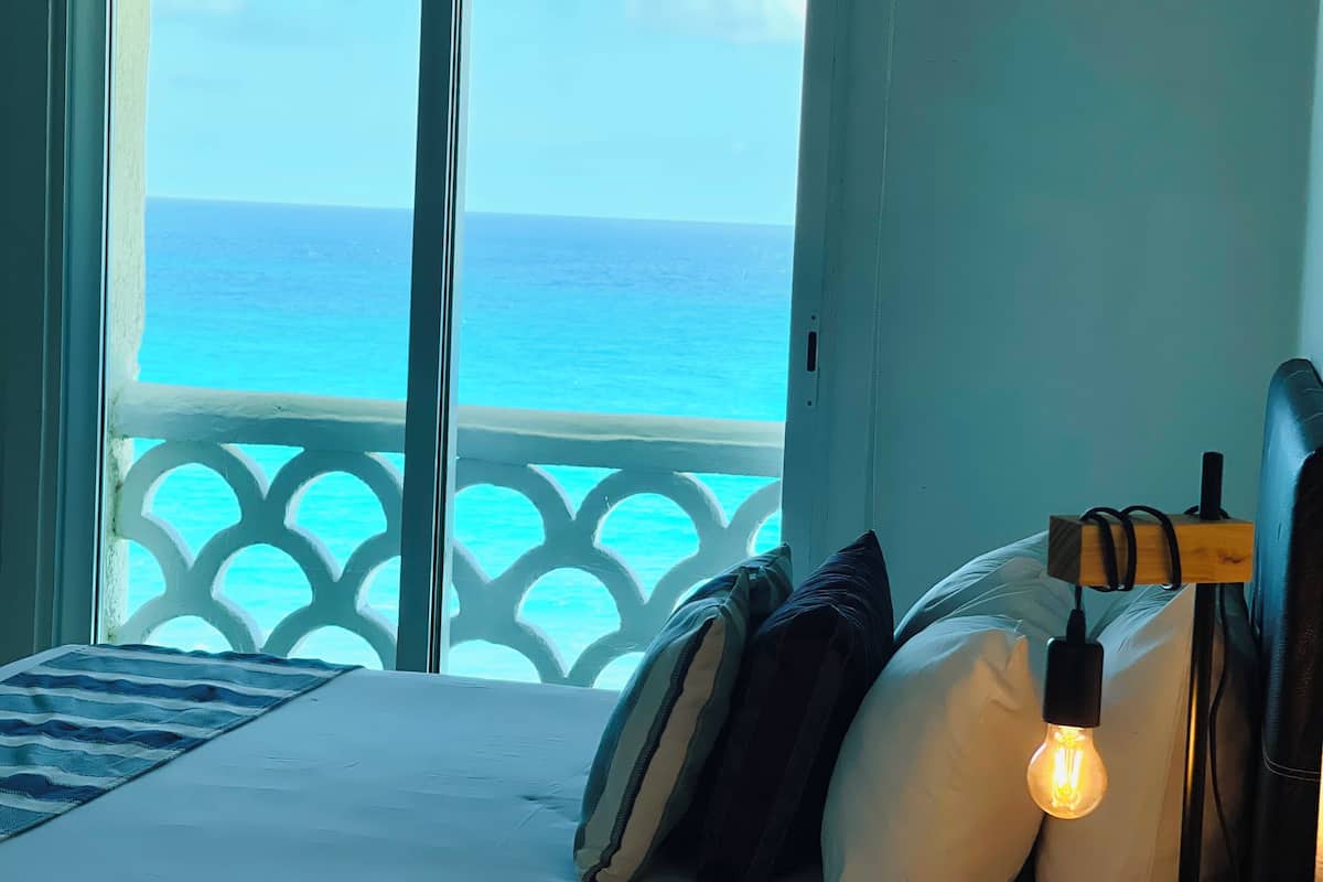 Ein Schlafzimmer mit Bett, Kissen, Nachttisch und einer Nachttischlampe. Ein Fenster bietet Blick auf den Ozean.