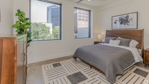 A modern bedroom with a large bed, two windows, a ceiling fan, and a view of buildings outside. - The Beacon Loft / Modern 1BR Downtown Apartment (Tulsa)