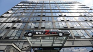 A high-rise building with a large glass facade and a car suspended in front of it. - Z Hotel New York (Long Island City)