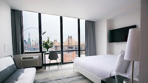 A hotel room with a large window, a bed, a sofa, a desk, and a TV. - Z Hotel New York (Long Island City)
