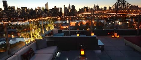 A rooftop terrace with a city skyline view, outdoor seating, and string lights.