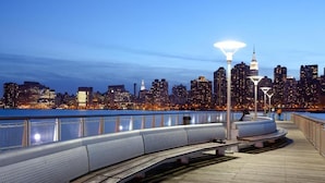 A waterfront promenade with a city skyline in the background, featuring modern buildings and a prominent tower. - Z Hotel New York (Long Island City)