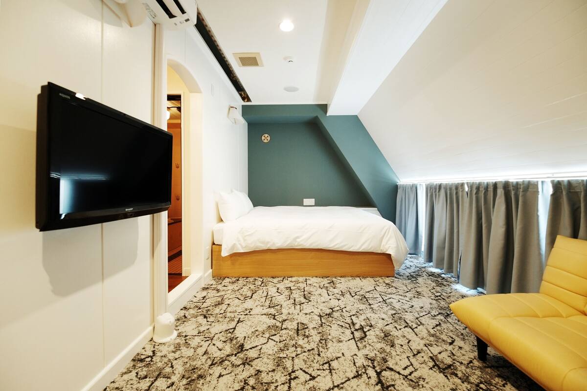 Een slaapkamer met een bed, een flatscreen-tv aan de muur, een kamerbreed tapijt met dessin en een gele fauteuil.