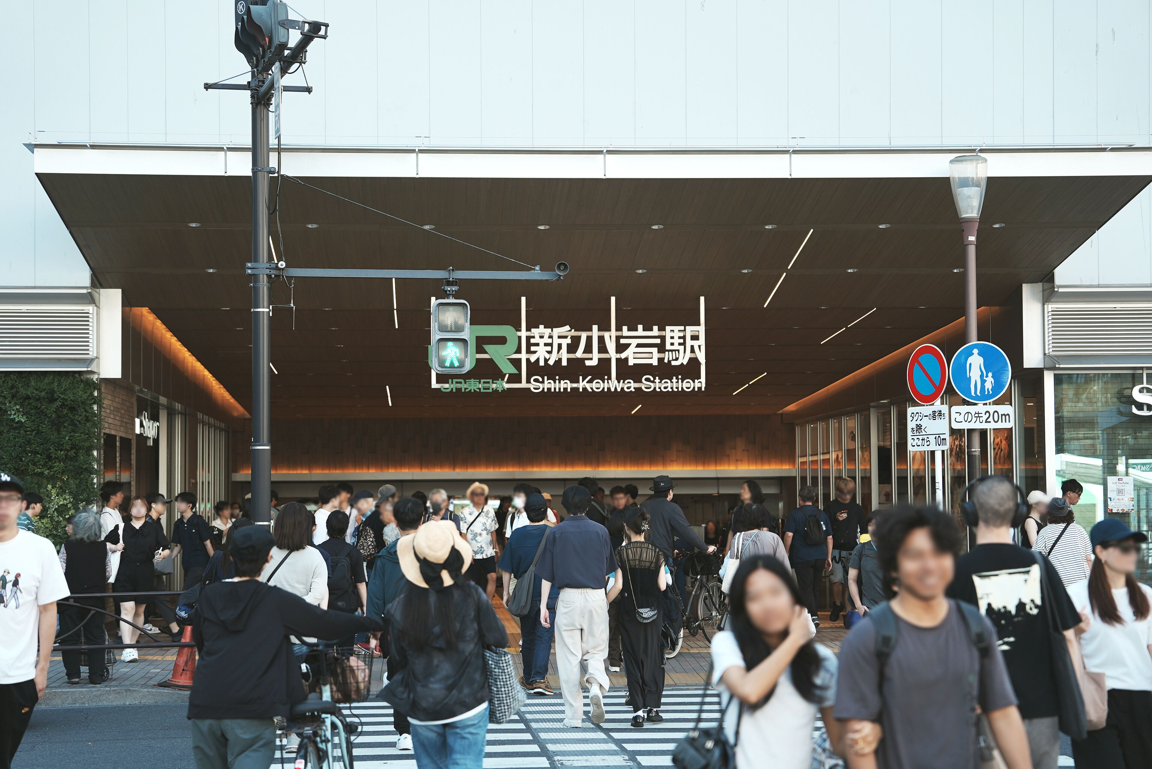 A crowded station entrance with a modern architectural design, a pedestrian crossing, and directional signs.