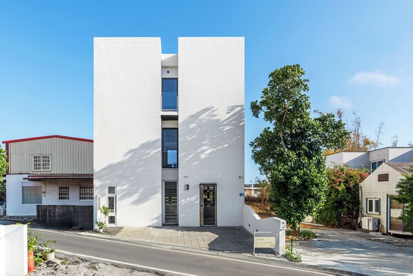A modern two-story building with a white facade, a central entrance, and a small garden area.