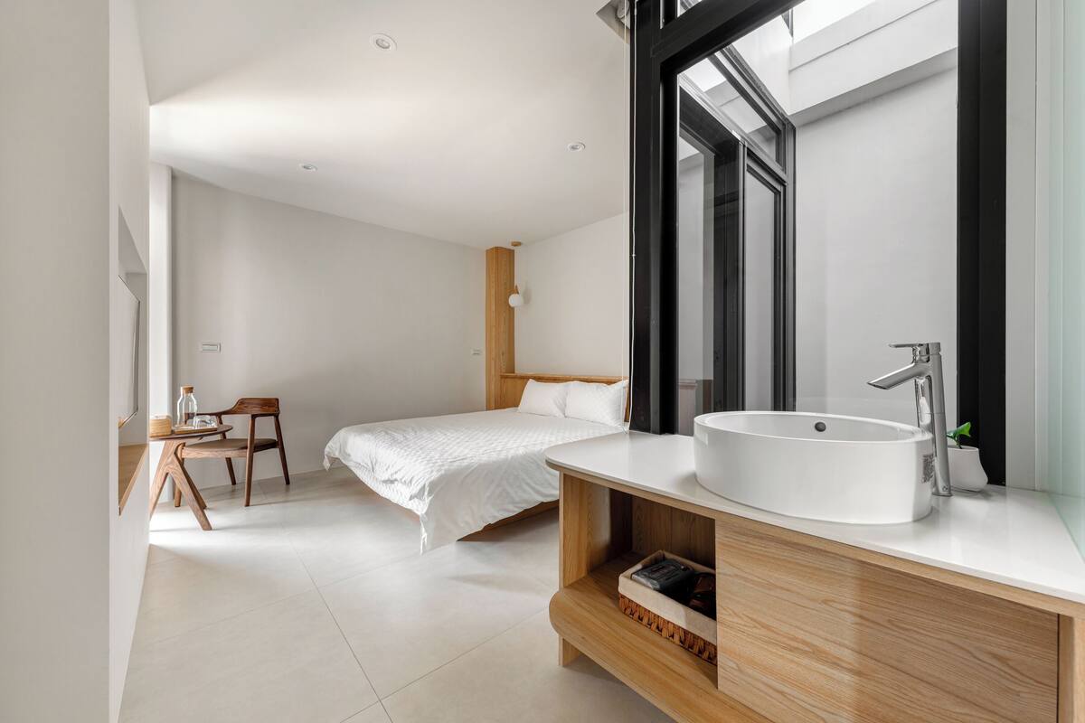 A modern bedroom with a large bed, a wooden nightstand, a white sink, and a large window.