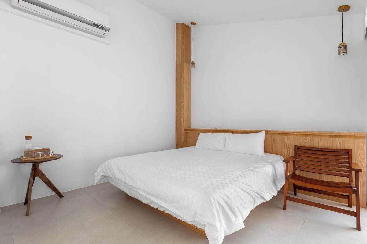 A bedroom with a bed, a wooden headboard, a small wooden table, and a wall-mounted air conditioner.