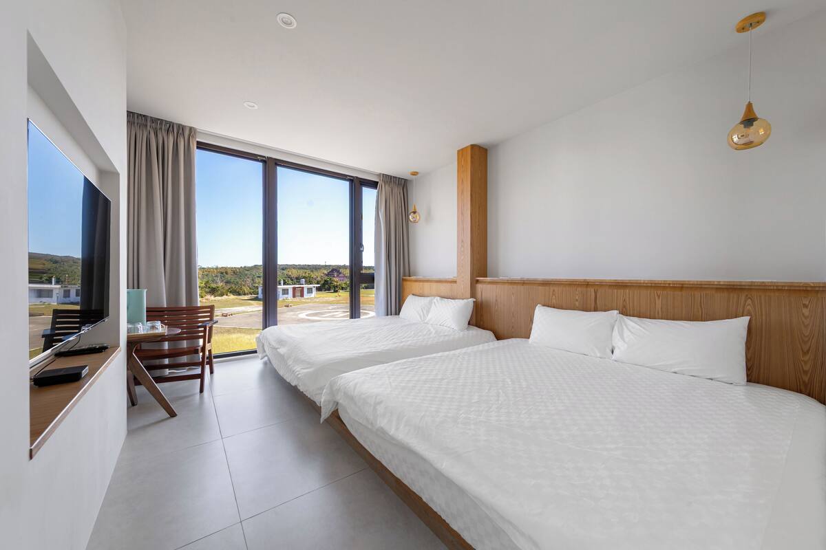 A hotel room with two beds, a desk with a chair, a TV, and large windows.