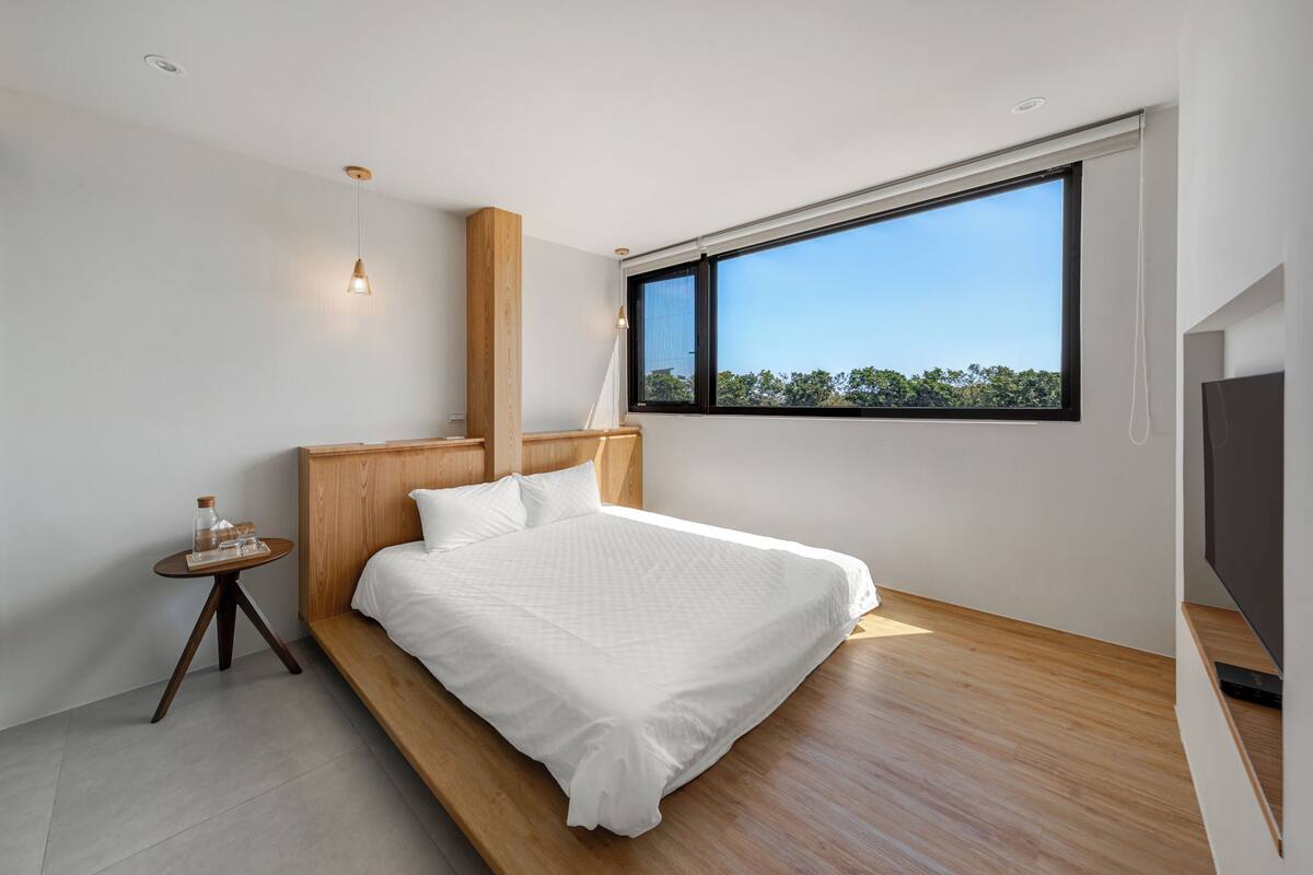 A modern bedroom with a large window, a bed, a bedside table, and a TV.