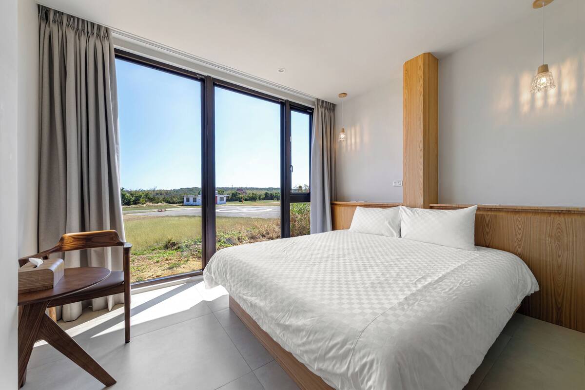 A bedroom with a large bed, a wooden headboard, a desk with a chair, and a view of a green landscape through large windows.