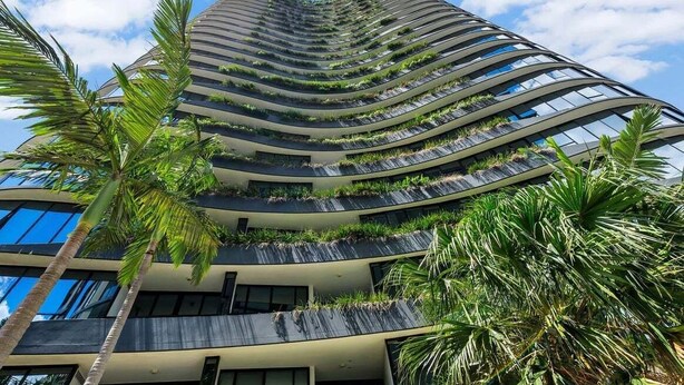 Un moderno edificio de gran altura con múltiples balcones, algunos con plantas, y palmeras alrededor.
