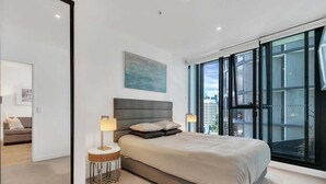 Un dormitorio moderno con una cama grande, una mesita de noche, una lámpara de pared y vistas a la ciudad a través de ventanales de gran altura.
