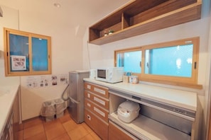 A compact kitchen with a microwave, a small refrigerator, and a sink. There are wooden shelves above the counter and a window with a view.