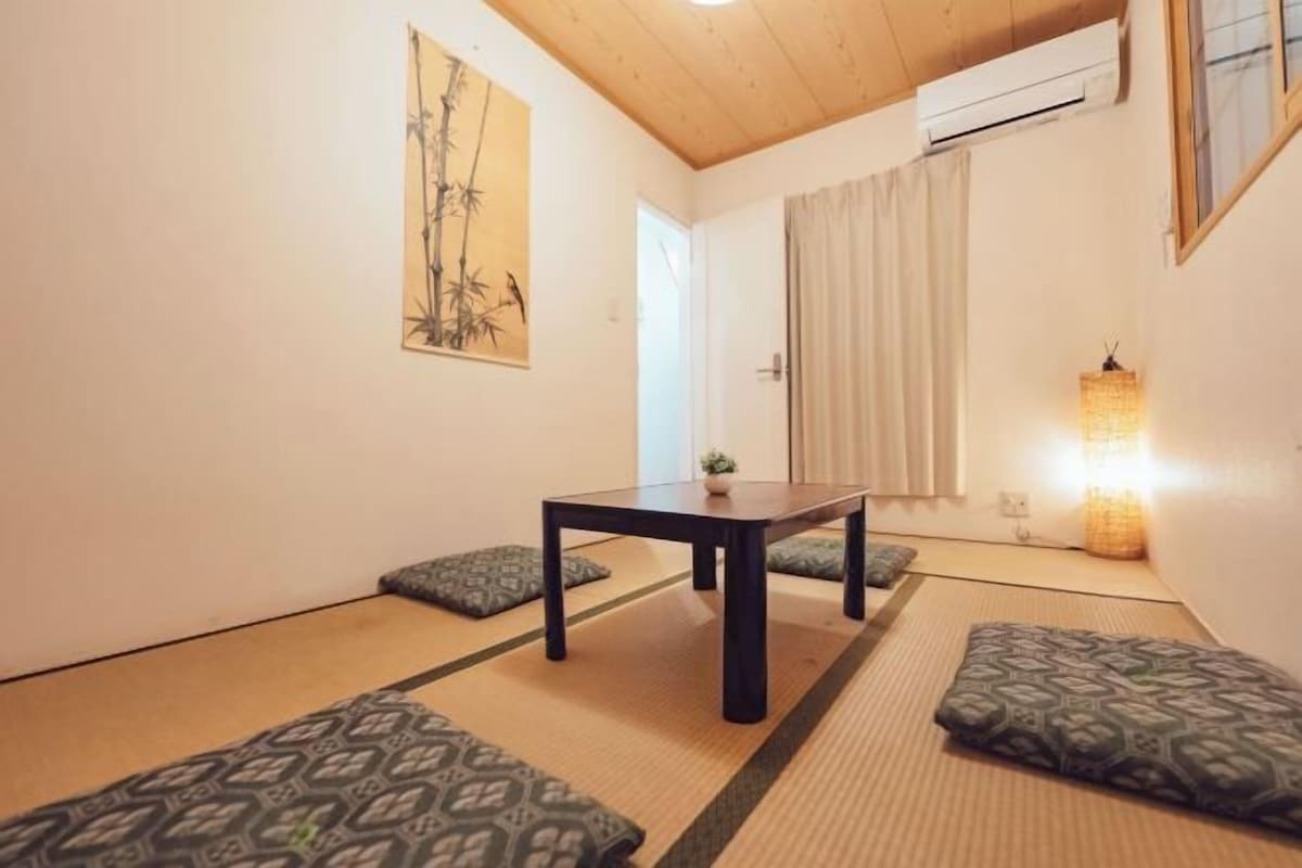 A traditional tatami mat room with a low table, cushions, a painting, and a lamp.