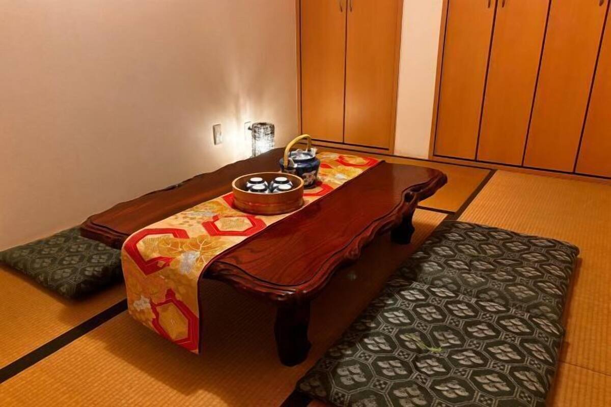 Una habitación japonesa tradicional con suelo de tatami, una mesa baja con camino de mesa tejido, un banco de madera y un juego de armarios de madera.