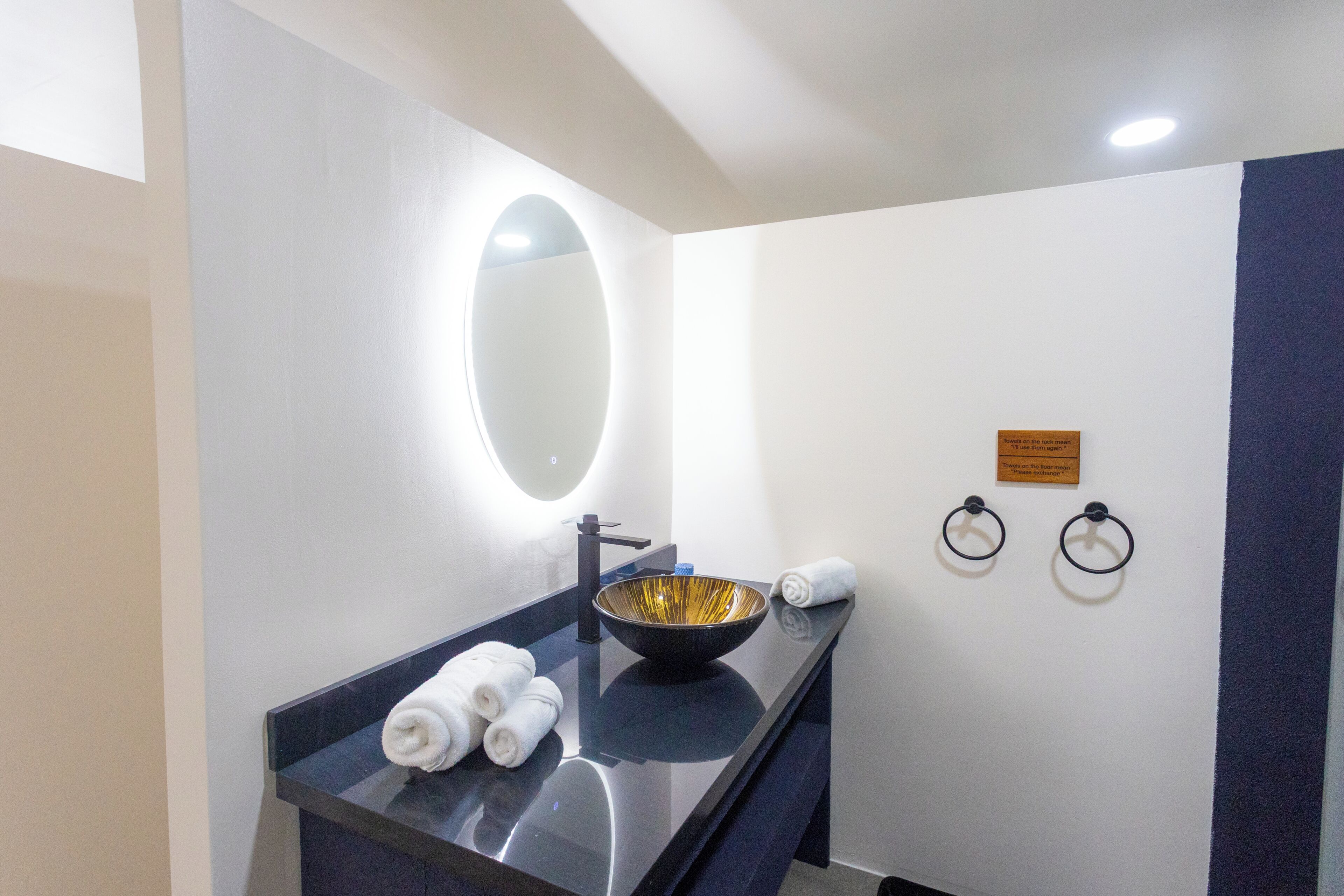 A modern bathroom with a black countertop, a gold bowl sink, a round mirror, and two towel rings.