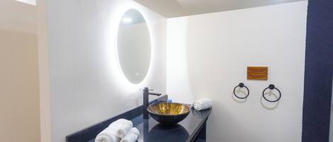 A modern bathroom with a black countertop, a gold bowl sink, a round mirror, and two towel rings.