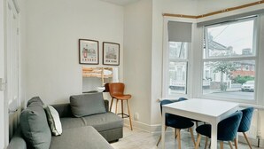 A modern living room with a grey sectional sofa, a dining table with chairs, and a large window with a view of buildings.
