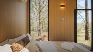 Un dormitorio con cama, almohadas, una lámpara fijada a la pared y vistas a árboles a través de un ventanal.