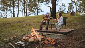 Two people seated at a table with a fire in front of them, surrounded by trees. - Three-Kalm by Tiny Away (Rainbow Flat)