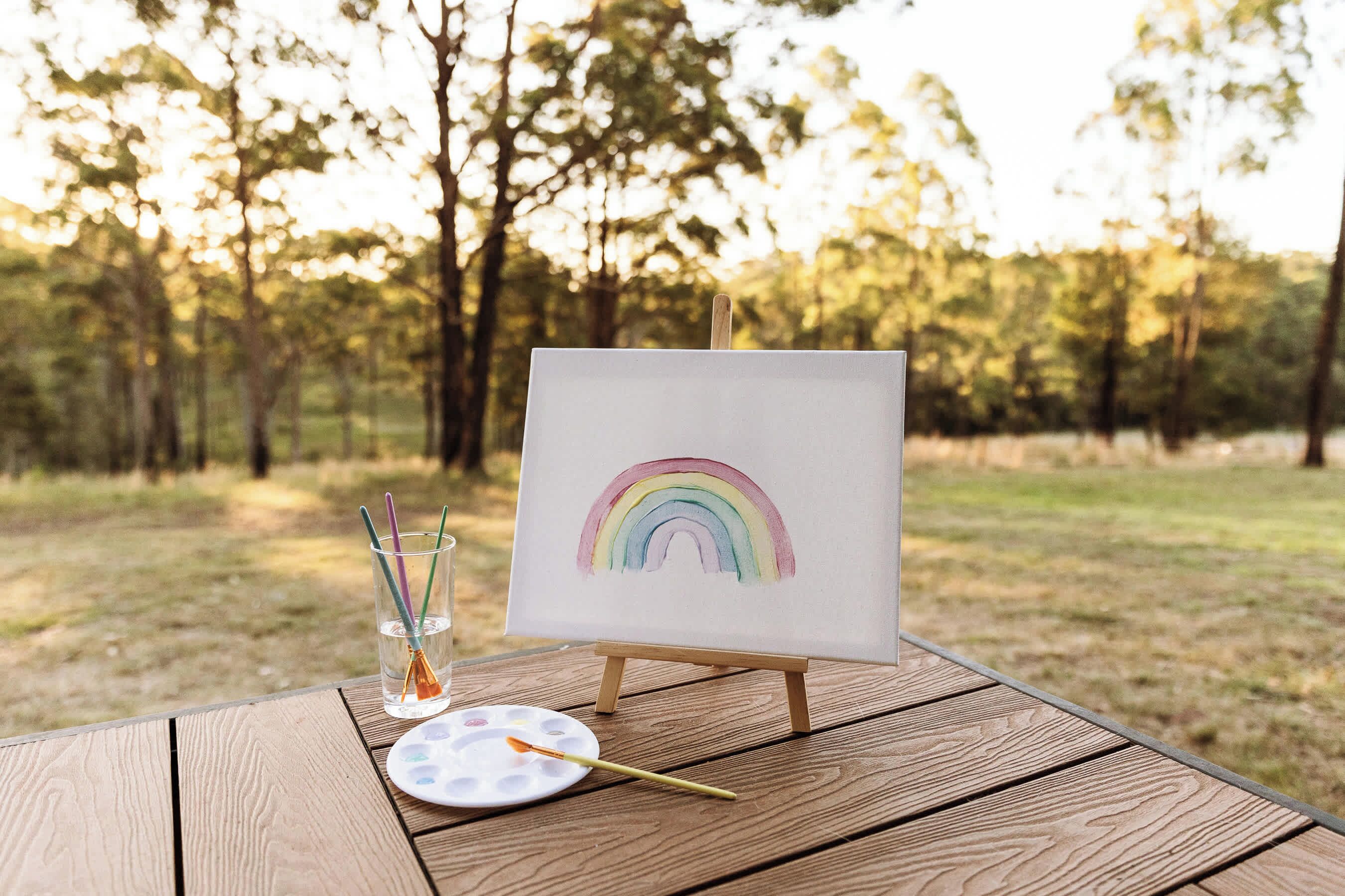 Un chevalet sur lequel est posé un tableau représentant un arc-en-ciel, le tout installé sur une table en bois, dans une forêt.