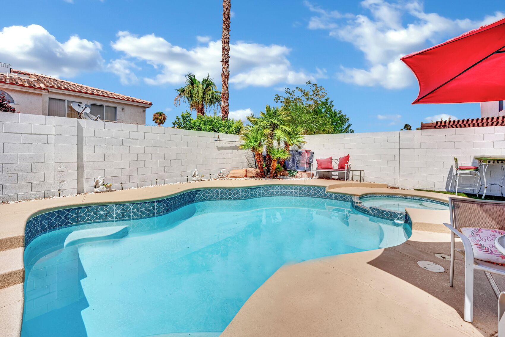 A swimming pool with a tiled edge, a red umbrella, and a patio area with chairs and a table.